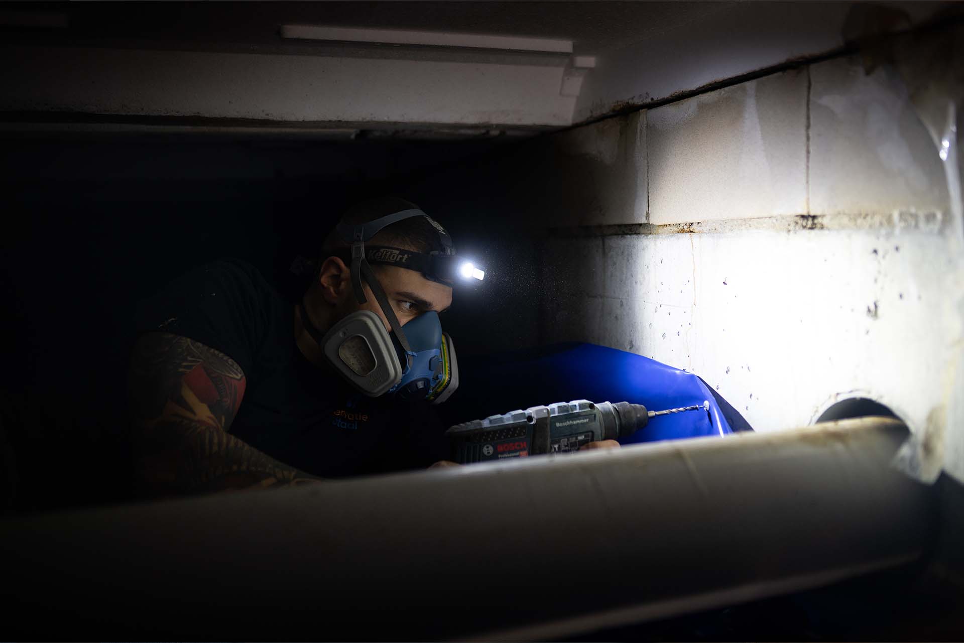 Medewerker met hoofdlamp en mondkapje aan het werk in een kruipruimte
